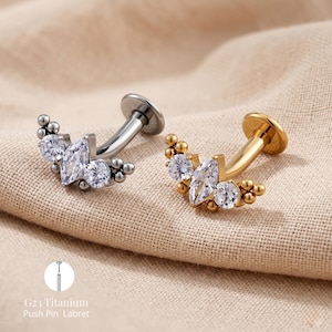 May include: Two labret studs with a curved post and flat back. One is silver-toned, the other gold-toned. Each features a cluster of clear, sparkling gemstones, including marquise and round cuts, and small bead accents. The text "G23 Titanium Push Pin Labret" is visible.