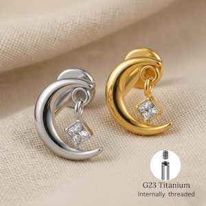 May include: Two crescent moon earrings, one silver and one gold, each with a dangling square-cut clear gemstone. The earrings have a post and are internally threaded. The text "G23 Titanium" and "Internally threaded" is also visible.