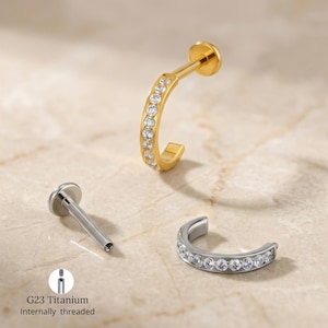 Op de afbeelding: Drie lichaamsjuwelen. Eén is een goudkleurige gebogen barbell met heldere edelstenen. Een andere is een zilverkleurige gebogen barbell met heldere edelstenen. Het derde stuk is een zilverkleurige labret stud. De tekst "G23 Titanium Internally threaded" is zichtbaar.
