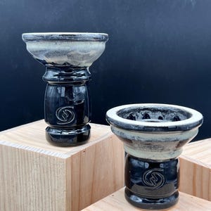 May include: Two ceramic hookah bowls. The bowls have a black base with a white and gray top. The base has a circular design. The bowls are displayed on top of wooden blocks against a dark background.