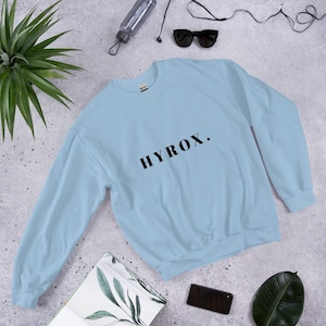 May include: A light blue sweatshirt with the word "HYROX" printed in black. The sweatshirt is made of a soft material and has a crew neck. The image also includes sunglasses, a water bottle, and a smartphone.