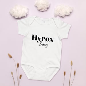 May include: White baby bodysuit featuring the text "Hyrox Baby" in black. The short-sleeved onesie has a classic design. The background is a soft purple colour with decorative elements.