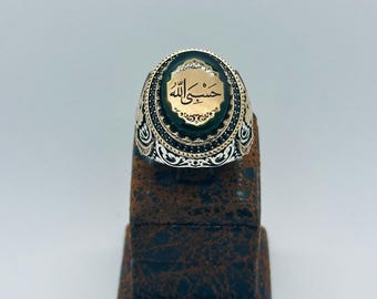 May include: A silver ring with a gold-colored inscription in Arabic script. The ring has a dark green center stone surrounded by black stones and ornate silver detailing. The ring is displayed on a brown, textured stand.