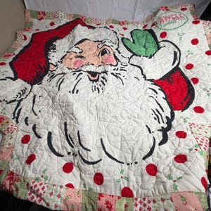 May include: A quilted blanket featuring a large graphic of a winking Santa Claus with a white beard, red hat, and green mitten. The background is white with red polka dots and green holly accents. The text "SWELL CHRISTMAS" is visible.