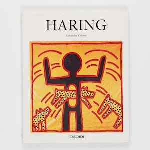 Puede incluir: Un libro de arte de tapa dura titulado HARING de Alexandra Kolossa, publicado por TASCHEN. La cubierta blanca presenta una figura humana negra central con el torso hueco, rodeada de criaturas amarillas y negras con manchas sobre un fondo naranja.