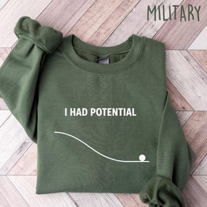 May include: Military green sweatshirt with the text "I HAD POTENTIAL" in white. The design includes a white line and a white ball. The sweatshirt is folded on a light wood-grain surface.