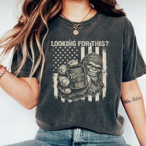 May include: Dark gray t-shirt with a graphic of a Bigfoot holding a 10mm object in front of an American flag. The text "LOOKING FOR THIS?" is above the graphic. The shirt is worn by a person wearing blue jeans and white shoes.