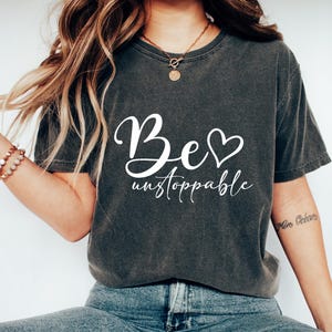 May include: Dark gray t-shirt with the words "Be unstoppable" in white script. The word "Be" has a heart replacing the "o". The shirt is paired with blue jeans and white slip-on shoes. A gold necklace and bracelets are also visible.