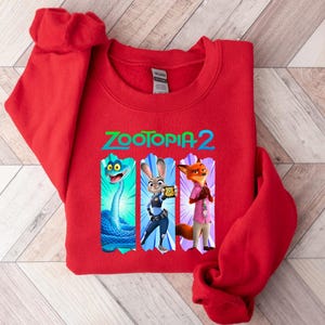 Puede incluir: Una sudadera roja con el texto "ZOOTOPIA 2" en verde y azul. El dise&ntilde;o incluye personajes de la pel&iacute;cula, como una serpiente azul, Judy Hopps y Nick Wilde. La sudadera est&aacute; hecha de un material suave.