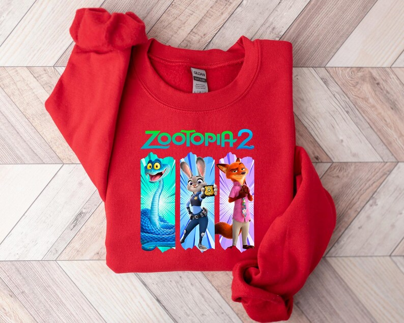Puede incluir: Una sudadera roja con el texto "ZOOTOPIA 2" en verde y azul. El dise&ntilde;o incluye personajes de la pel&iacute;cula, como una serpiente azul, Judy Hopps y Nick Wilde. La sudadera est&aacute; hecha de un material suave.