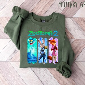 Puede incluir: Sudadera verde militar con el texto "Zootopia 2" y tres ilustraciones de personajes de dibujos animados: una serpiente, un conejo y un zorro. La sudadera est&aacute; doblada sobre una superficie de vetas de madera clara.