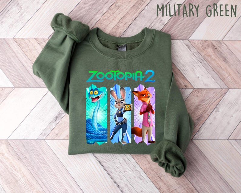 Puede incluir: Sudadera verde militar con el texto "Zootopia 2" y tres ilustraciones de personajes de dibujos animados: una serpiente, un conejo y un zorro. La sudadera est&aacute; doblada sobre una superficie de vetas de madera clara.