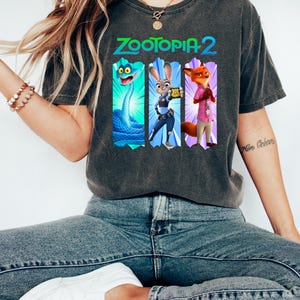 Puede incluir: Camiseta gris oscuro con el texto "ZOOTOPIA 2" e ilustraciones de personajes. Las ilustraciones incluyen una serpiente azul, un conejo con uniforme de polic&iacute;a y un zorro con una camisa rosa. La camiseta la lleva una persona con vaqueros azules y zapatos blancos.