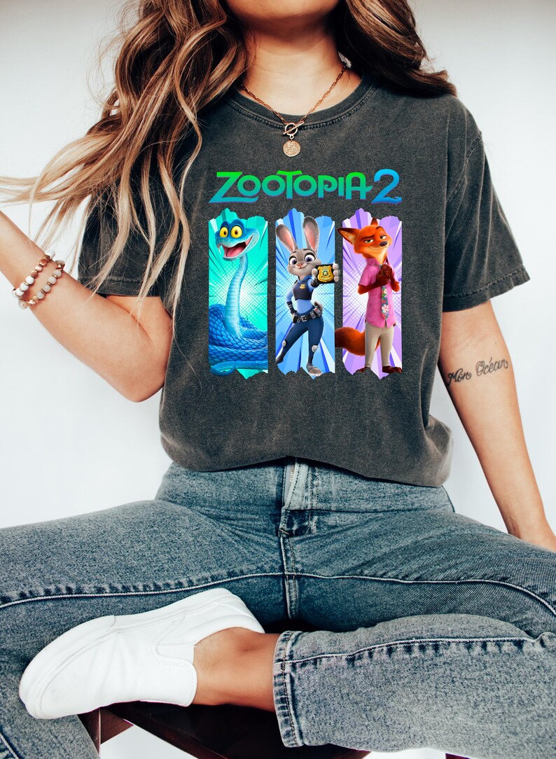 Puede incluir: Camiseta gris oscuro con el texto "ZOOTOPIA 2" e ilustraciones de personajes. Las ilustraciones incluyen una serpiente azul, un conejo con uniforme de polic&iacute;a y un zorro con una camisa rosa. La camiseta la lleva una persona con vaqueros azules y zapatos blancos.