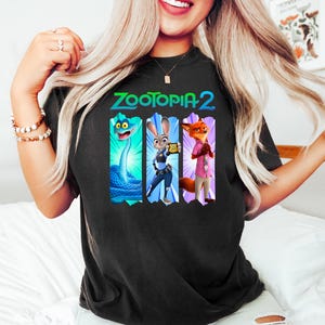 Puede incluir: Camiseta negra con el texto "ZOOTOPIA 2" y tres personajes de dibujos animados. Los personajes son una serpiente azul, un conejo con un dispositivo y un zorro con una camisa rosa. La camiseta es de color oscuro.