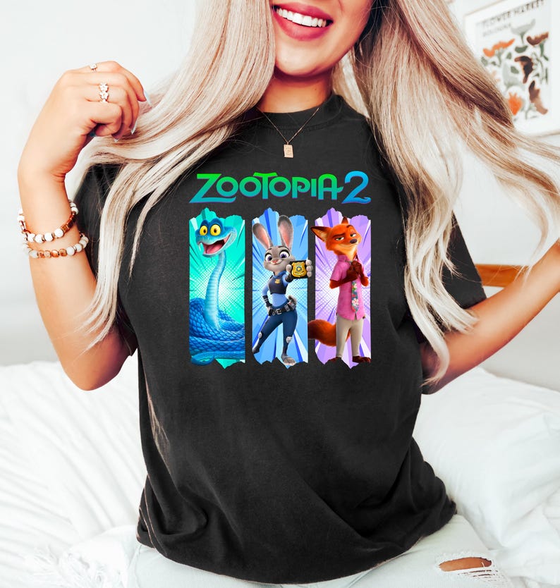Puede incluir: Camiseta negra con el texto "ZOOTOPIA 2" y tres personajes de dibujos animados. Los personajes son una serpiente azul, un conejo con un dispositivo y un zorro con una camisa rosa. La camiseta es de color oscuro.