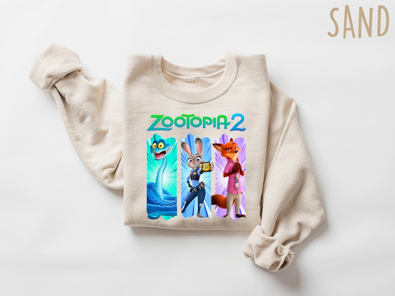 Puede incluir: Una sudadera beige con el texto "ZOOTOPIA 2" en verde y azul. Debajo, tres paneles verticales con personajes de dibujos animados: una serpiente azul, un conejo con uniforme de polic&iacute;a y un zorro con camisa rosa.