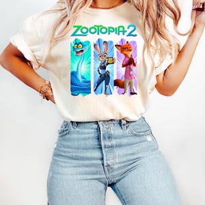 Puede incluir: Camiseta color crema con el texto "ZOOTOPIA 2" en verde y azul. Debajo, tres paneles verticales con personajes de dibujos animados: una serpiente azul, un conejo con uniforme de polic&iacute;a y un zorro con camisa rosa.