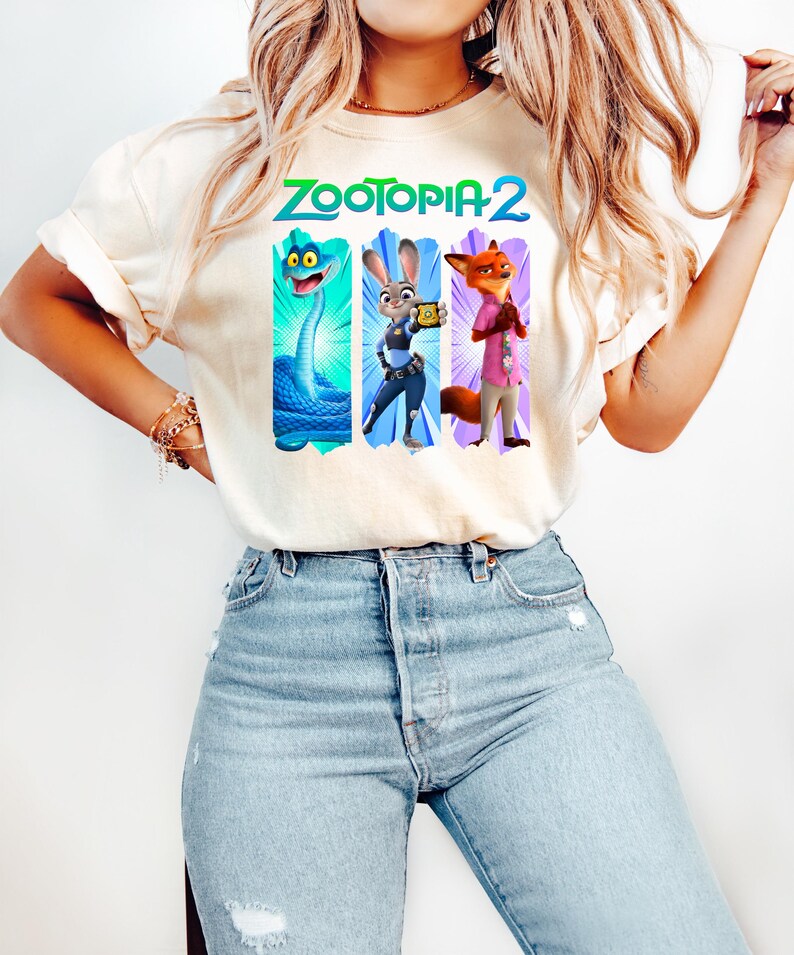 Puede incluir: Camiseta color crema con el texto "ZOOTOPIA 2" en verde y azul. Debajo, tres paneles verticales con personajes de dibujos animados: una serpiente azul, un conejo con uniforme de polic&iacute;a y un zorro con camisa rosa.