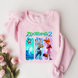 Puede incluir: Una sudadera rosa claro con el texto "ZOOTOPIA 2" en verde y azul. El dise&ntilde;o incluye tres personajes de dibujos animados: una serpiente azul, un conejo y un zorro. La sudadera es de material suave.