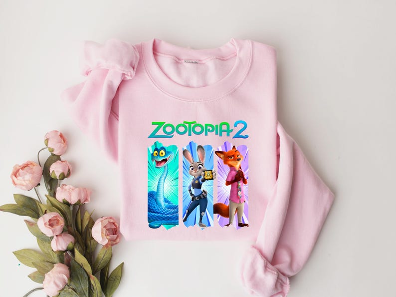 Puede incluir: Una sudadera rosa claro con el texto "ZOOTOPIA 2" en verde y azul. El dise&ntilde;o incluye tres personajes de dibujos animados: una serpiente azul, un conejo y un zorro. La sudadera es de material suave.