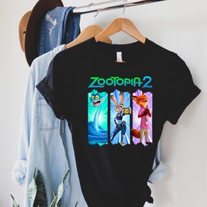 Puede incluir: Camiseta negra con el texto "ZOOTOPIA 2" en verde y azul. El dise&ntilde;o presenta tres personajes de dibujos animados: una serpiente azul, un conejo con uniforme de polic&iacute;a y un zorro con camisa rosa. La camiseta est&aacute; colgada en una percha de madera.