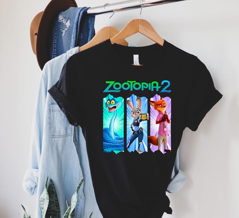 Puede incluir: Camiseta negra con el texto "ZOOTOPIA 2" en verde y azul. El dise&ntilde;o presenta tres personajes de dibujos animados: una serpiente azul, un conejo con uniforme de polic&iacute;a y un zorro con camisa rosa. La camiseta est&aacute; colgada en una percha de madera.