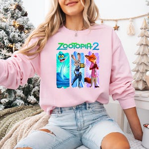 Puede incluir: Una sudadera rosa claro con el texto "ZOOTOPIA 2" en verde y azul. El dise&ntilde;o presenta tres personajes de dibujos animados: una serpiente azul, un conejo con uniforme de polic&iacute;a y un zorro con camisa rosa. La sudadera tiene mangas largas.