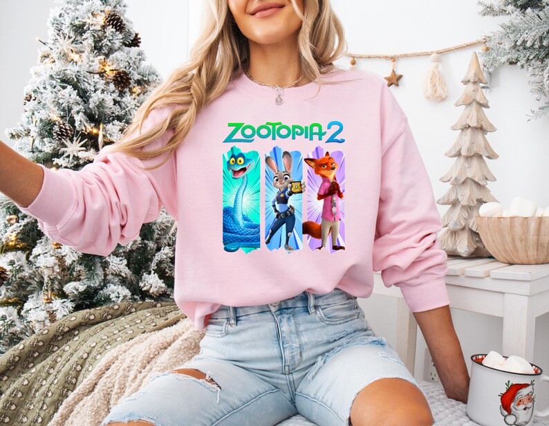 Puede incluir: Una sudadera rosa claro con el texto "ZOOTOPIA 2" en verde y azul. El dise&ntilde;o presenta tres personajes de dibujos animados: una serpiente azul, un conejo con uniforme de polic&iacute;a y un zorro con camisa rosa. La sudadera tiene mangas largas.