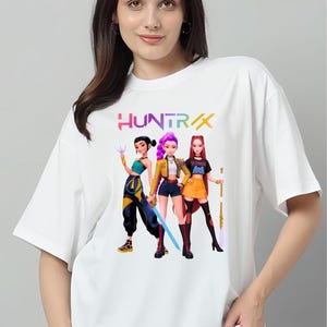 May include: White t-shirt with a vibrant graphic of three animated female characters, each holding a weapon. The word "HUNTRAX" is printed above the characters in a multi-coloured font. The design is centered on the shirt.