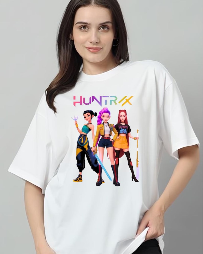 May include: White t-shirt with a vibrant graphic of three animated female characters, each holding a weapon. The word "HUNTRAX" is printed above the characters in a multi-coloured font. The design is centered on the shirt.