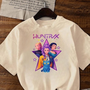 May include: Cream-coloured t-shirt with the word "HUNTRAX" above a graphic of three women and a blue cat inside a star. The women have different hair colours and wear various outfits. The shirt has short sleeves.