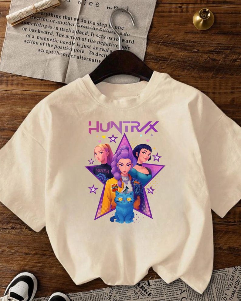 May include: Cream-coloured t-shirt with the word "HUNTRAX" above a graphic of three women and a blue cat inside a star. The women have different hair colours and wear various outfits. The shirt has short sleeves.