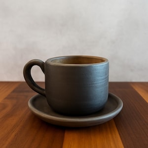 May include: A dark grey ceramic coffee cup and saucer set. The cup features a rounded handle and a textured finish. The interior of the cup is a lighter brown. The set is displayed on a wooden surface.