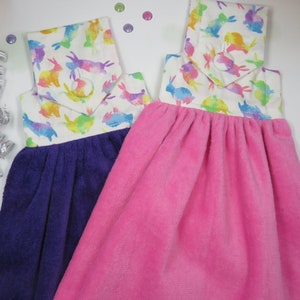 May include: Two hanging kitchen towels. One is purple, the other is pink. Both have a white top with a colorful bunny pattern. The towels are made of a soft, absorbent material and are designed to hang from a hook.