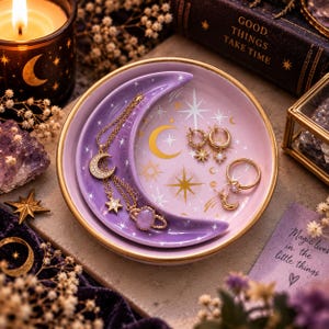 Könnte beinhalten: Eine runde Schmuckschale aus Keramik mit einer mondsichelförmigen Vertiefung, verziert mit einem hellvioletten und lavendelfarbenen Himmelsdesign mit goldenen Sternen und Monden. Die Schale enthält eine goldene Kette, Ohrringe und einen Ring.