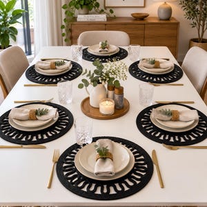 May include: A dining table set for six with white plates, beige linen napkins in woven rings, and black circular placemats with a scalloped edge. Gold-toned cutlery, clear textured glasses, and a central wooden tray with a white pitcher and candle.