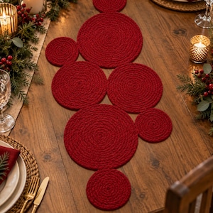 May include: A table runner composed of multiple circular, deep red, braided rope mats of varying sizes arranged in a cluster. The textured, coiled design creates a continuous decorative centerpiece that lies flat across the wooden surface.