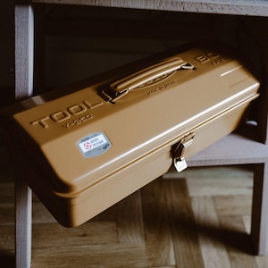 Op de afbeelding: Een bruin metalen gereedschapskist met een handvat, sluiting en de tekst "TOOL Y-350". De gereedschapskist heeft een "GOOD DESIGN" sticker en de tekst "MADE IN JAPAN". De gereedschapskist ligt op een houten plank.