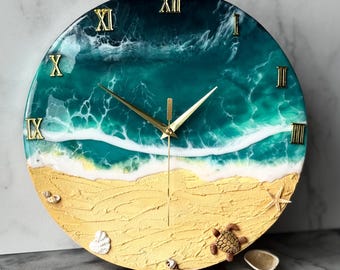 May include: Round wall clock with a beach scene design. The clock face features a sandy beach, ocean waves, and gold Roman numerals. Decorative starfish, seashells, and a turtle are scattered on the clock face. The clock hands are gold.