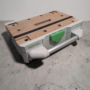 May include: A Festool Systainer storage box, featuring a light brown wooden top with black stripes and multiple holes. The box has a white body with a green latch and a silver handle. The brand name "Festool" is printed in green on the side.