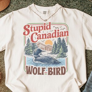 May include: Cream-colored t-shirt with a graphic that reads "Stupid Canadian Wolf Bird." The graphic features a loon on a lake with trees and a sunset. The shirt is styled with a necklace, shoes, and a hat.