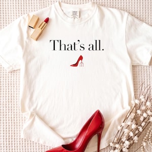 May include: A white t-shirt with the text "That's all." and a red high heel graphic. A red stiletto heel and lipstick are next to the shirt. A pearl necklace and decorative flowers are also in the image.