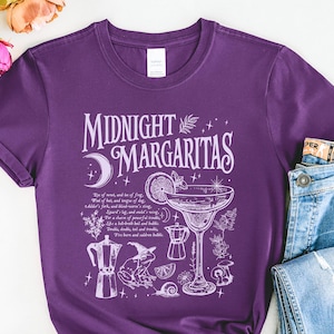 May include: A purple t-shirt with the words "Midnight Margaritas" in white script, along with a cocktail illustration and other botanical elements. The shirt is paired with blue jeans.