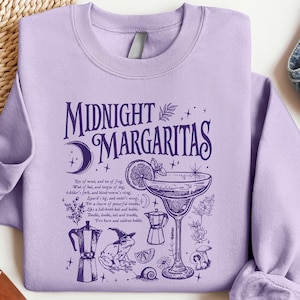 May include: A light purple sweatshirt with the words "Midnight Margaritas" in dark purple. The graphic includes a margarita, a frog wearing a witch hat, and other botanical elements. The sweatshirt is displayed with sunglasses, shoes, and jeans.