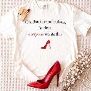 May include: White t-shirt with the text "Oh, don't be ridiculous, Andrea, everyone wants this" and a red high heel graphic. A red lipstick and a red high heel shoe are next to the shirt. A pearl necklace and white flowers are also in the image.