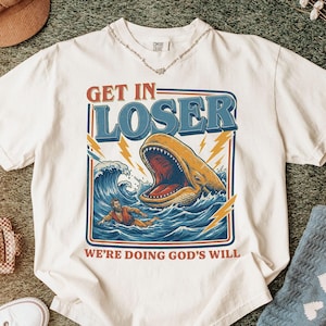 May include: White t-shirt with a graphic design featuring a whale with an open mouth in the ocean, a person in the water, and the text "GET IN LOSER WE'RE DOING GOD'S WILL" in a retro style.