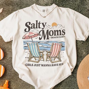 May include: Cream-colored t-shirt with the text "Salty Moms Beach Club" and a beach scene graphic. The graphic includes two beach chairs, an umbrella, and the phrase "Girls Just Wanna Have Sun."