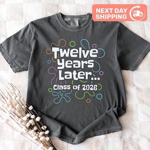May include: Dark gray t-shirt with the text "Twelve Years Later... Class of 2026" in white, with colorful decorative shapes. The shirt has a crew neck and short sleeves. A "Next Day Shipping" sticker is in the top right corner.
