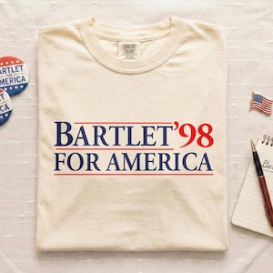 May include: Cream-colored t-shirt with the text "BARTLET '98 FOR AMERICA" in navy blue and red. Two round buttons and a small American flag pin are also visible. A notepad with the text "Bartlet '98" and a pen are on the right.